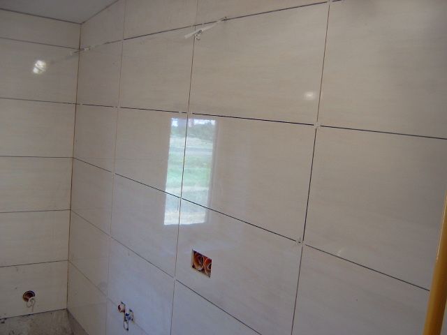 Ściana w trakcie kafelkowania jasnymi, błyszczącymi płytkami ceramicznymi, widoczne krzyżyki dystansowe, przygotowane otwory na instalacje elektryczne i wodno-kanalizacyjne.