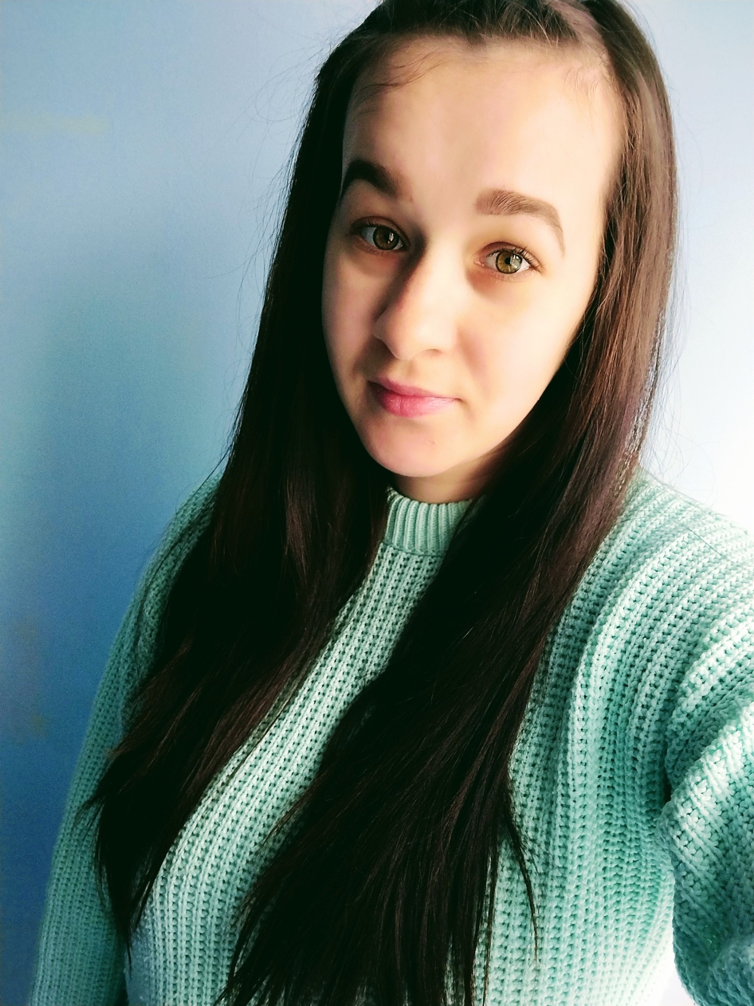 Portret młodej kobiety z długimi, ciemnymi włosami i zielonymi oczami, ubrana w miętowy sweter na tle jasnoniebieskiej ściany.