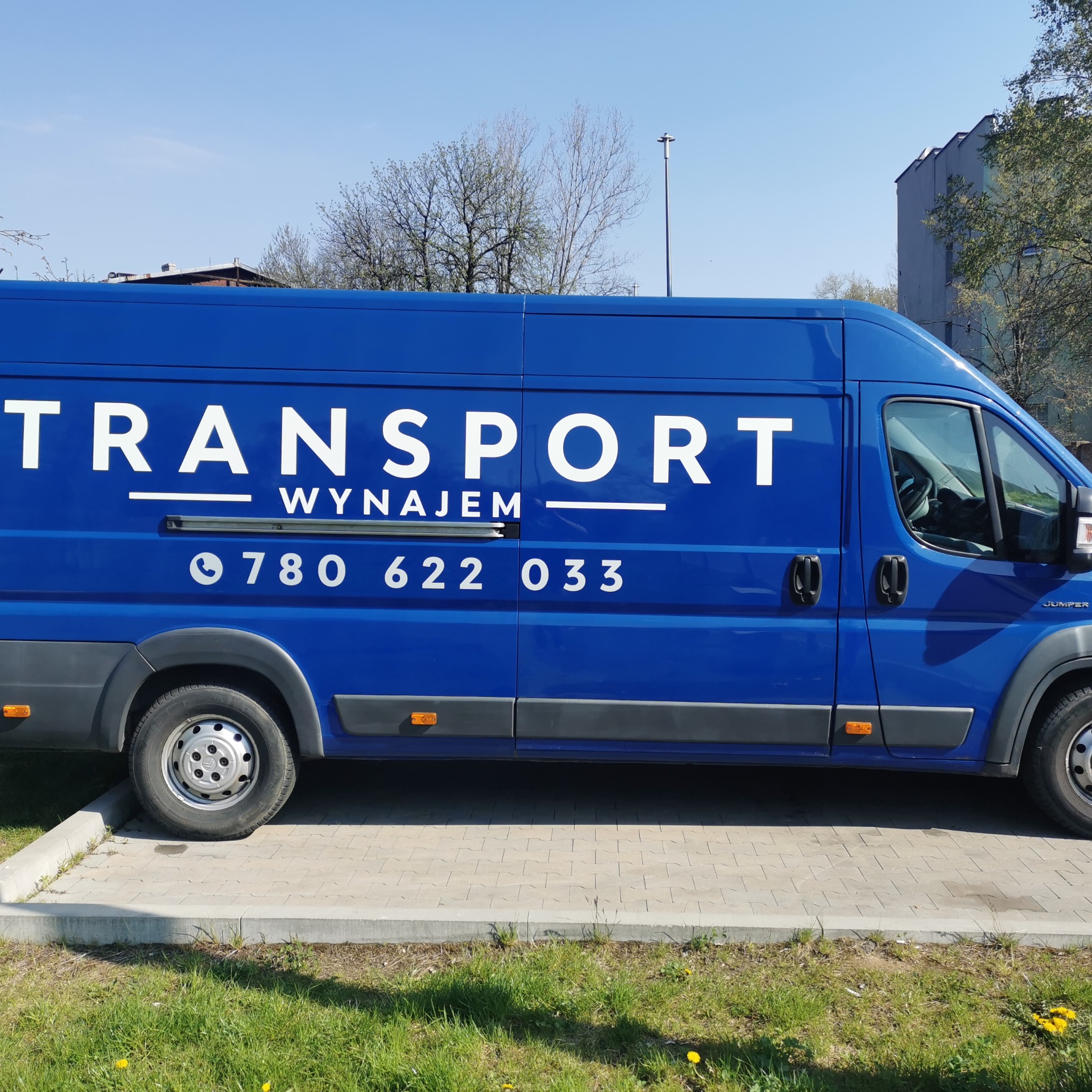 Niebieski samochód dostawczy z białym napisem 'TRANSPORT WYNAJEM' i numerem telefonu, zaparkowany na kostce brukowej obok trawnika z żółtymi kwiatami.