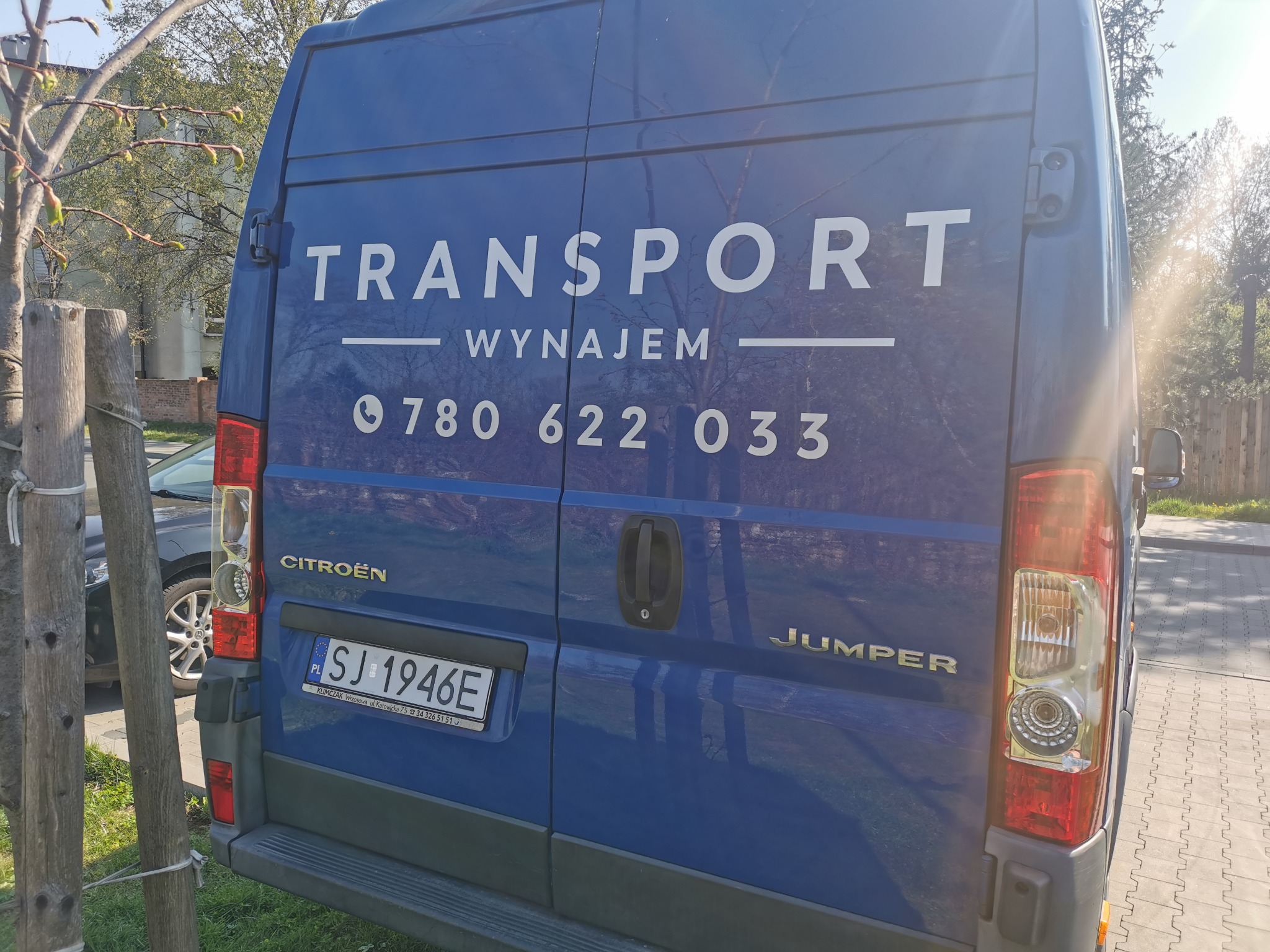 Tył niebieskiego samochodu dostawczego Citroen Jumper z napisem 'TRANSPORT WYNAJEM' i numerem telefonu, zaparkowanego na zewnątrz w słoneczny dzień.