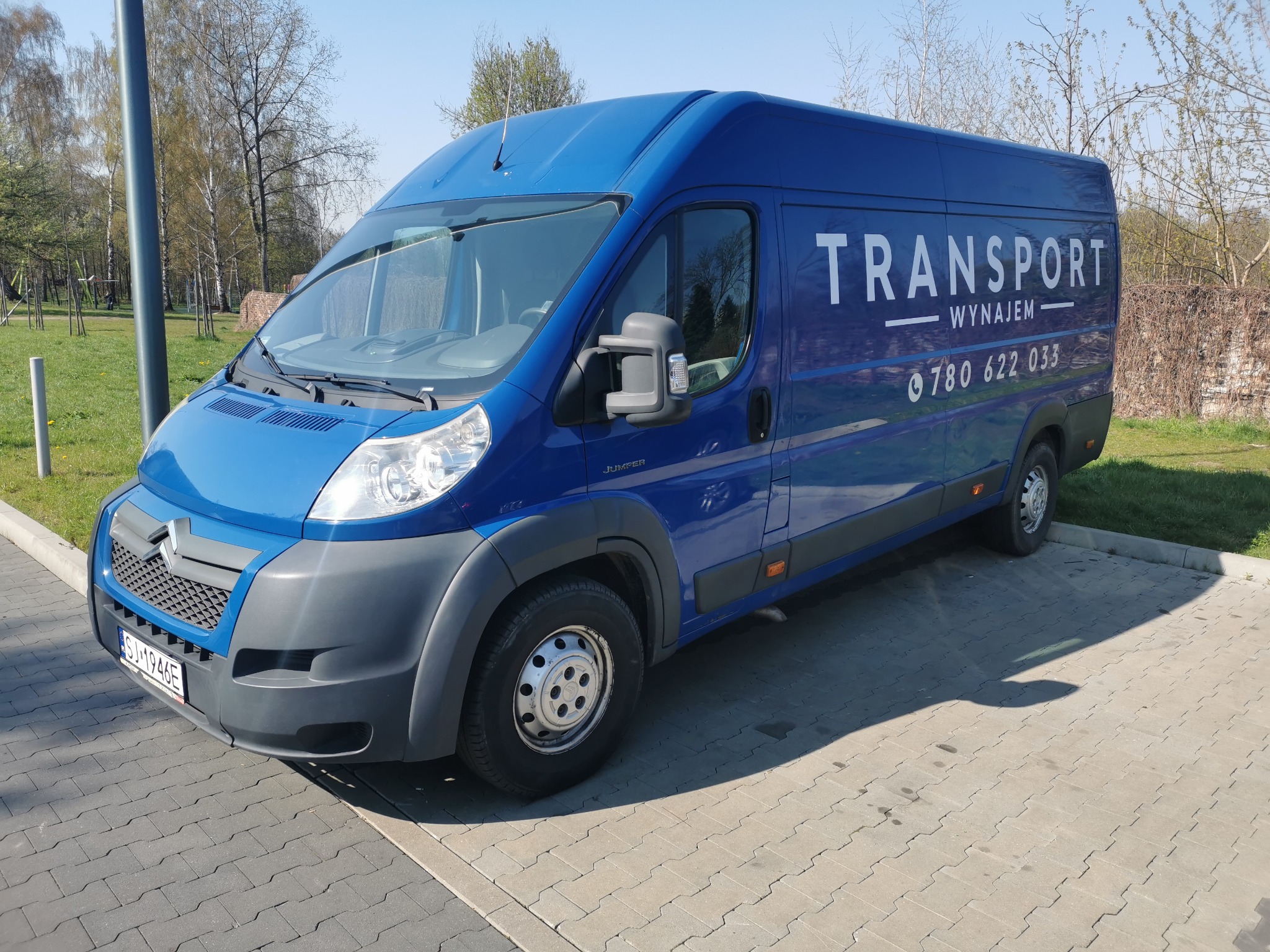 Niebieski samochód dostawczy z napisem 'TRANSPORT WYNAJEM' i numerem telefonu zaparkowany na kostce brukowej, z widoczną tablicą rejestracyjną i elementami zieleni w tle.