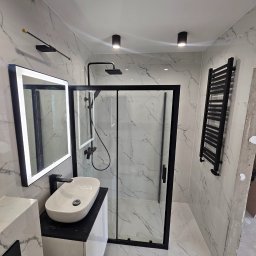 Marcin Szymczak - Nowoczesna łazienka z białymi marmurkowymi płytkami, czarną armaturą i kabiną prysznicową typu walk-in z czarnymi profilami, podświetlane lustro i czarny grzejnik drabinkowy, sufitowe lampy punktowe.