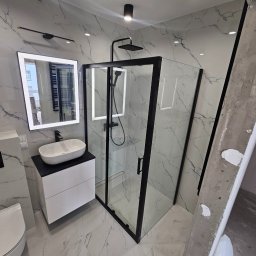 Marcin Szymczak - Nowoczesna łazienka z marmurowymi płytkami, czarną armaturą i kabiną prysznicową typu walk-in, widoczna drabina malarska z resztkami farby.