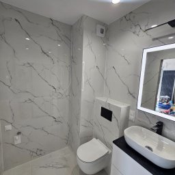 Marcin Szymczak - Nowoczesna łazienka z białymi płytkami imitującymi marmur, wisząca toaleta z czarnym panelem, owalna umywalka nablatowa na białej szafce z czarnym blatem i podświetlanym lustrem.