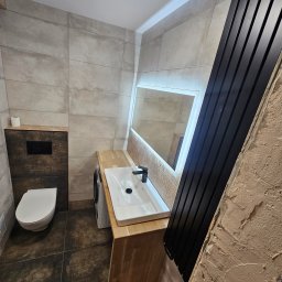 Marcin Szymczak - Nowoczesna łazienka z beżowymi płytkami, drewnianym blatem pod umywalką, podświetlanym lustrem i czarnym grzejnikiem drabinkowym, widoczna pralka i toaleta.