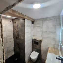 Nowoczesna łazienka w trakcie wykańczania: widoczna kabina prysznicowa z deszczownicą w kolorze miedzi, podwieszana toaleta, umywalka nablatowa na drewnianym blacie oraz fragment odsłoniętej ściany...