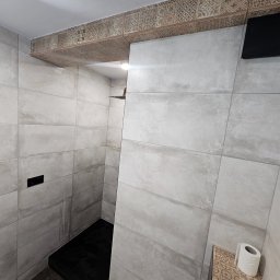 Marcin Szymczak - Nowoczesna łazienka z szarymi, prostokątnymi płytkami, czarnym brodzikiem i dekoracyjnym pasem płytek z geometrycznym wzorem pod sufitem oraz rolką papieru toaletowego na półce.