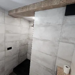 Nowoczesna łazienka z szarymi, prostokątnymi płytkami, czarnym brodzikiem i dekoracyjnym pasem płytek z geometrycznym wzorem pod sufitem oraz rolką papieru toaletowego na półce.