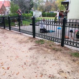Marcin Szymczak - Montaż ozdobnego, metalowego ogrodzenia posesji z okrągłymi elementami dekoracyjnymi; w tle dom jednorodzinny i osoba fotografująca postęp prac.