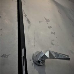 Marcin Szymczak - Szara ściana z efektem betonu i nowoczesna, geometryczna klamka. Widoczna czarna szczelina dylatacyjna. Minimalistyczny design wnętrza.