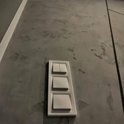 Marcin Szymczak - Szara ściana z dekoracyjnym tynkiem i potrójnym, białym włącznikiem światła. Minimalistyczne wykończenie wnętrza z bliska, z widoczną fakturą ściany.