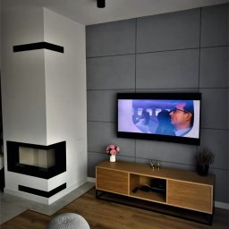 Marcin Szymczak - Nowoczesny salon z kominkiem narożnym, szarą ścianą z paneli i drewnianą szafką RTV. Na podłodze panele i pufa. Minimalistyczne oświetlenie sufitowe.