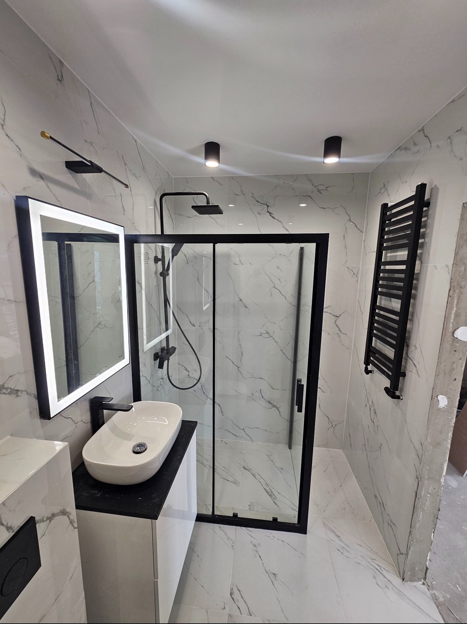 Nowoczesna łazienka z białymi marmurkowymi płytkami, czarną armaturą i kabiną prysznicową typu walk-in z czarnymi profilami, podświetlane lustro i czarny grzejnik drabinkowy, sufitowe lampy punktowe.