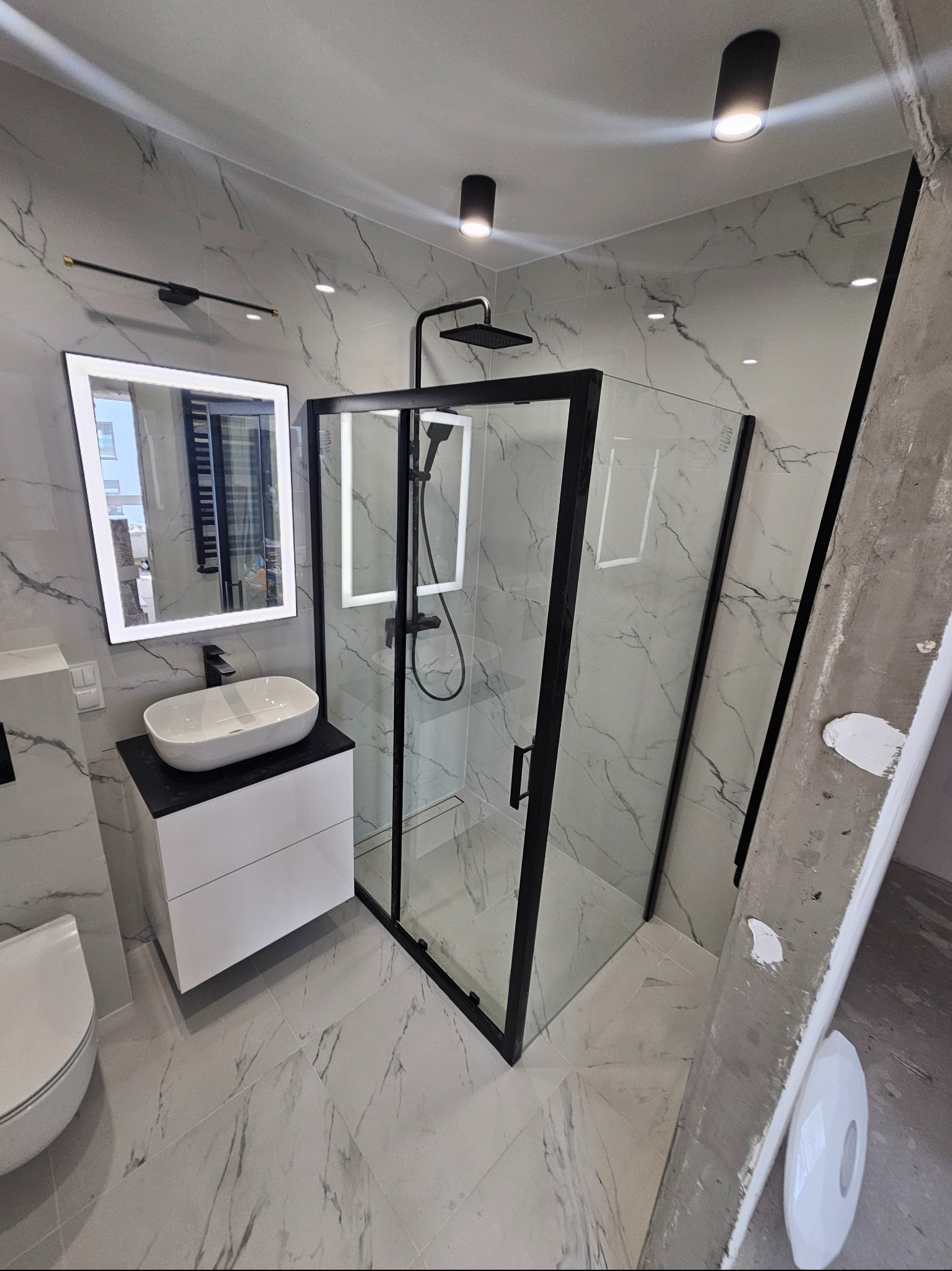 Nowoczesna łazienka z marmurowymi płytkami, czarną armaturą i kabiną prysznicową typu walk-in, widoczna drabina malarska z resztkami farby.