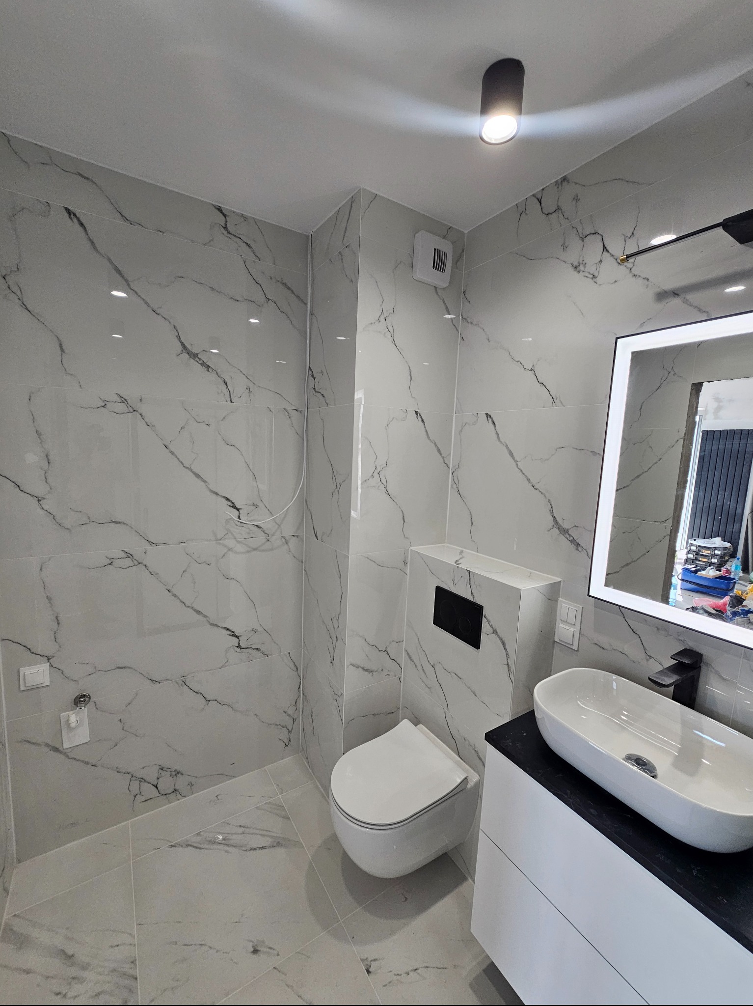 Nowoczesna łazienka z białymi płytkami imitującymi marmur, wisząca toaleta z czarnym panelem, owalna umywalka nablatowa na białej szafce z czarnym blatem i podświetlanym lustrem.