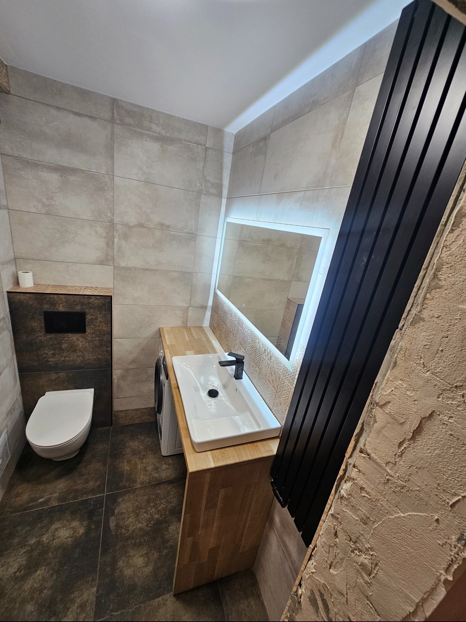 Nowoczesna łazienka z beżowymi płytkami, drewnianym blatem pod umywalką, podświetlanym lustrem i czarnym grzejnikiem drabinkowym, widoczna pralka i toaleta.