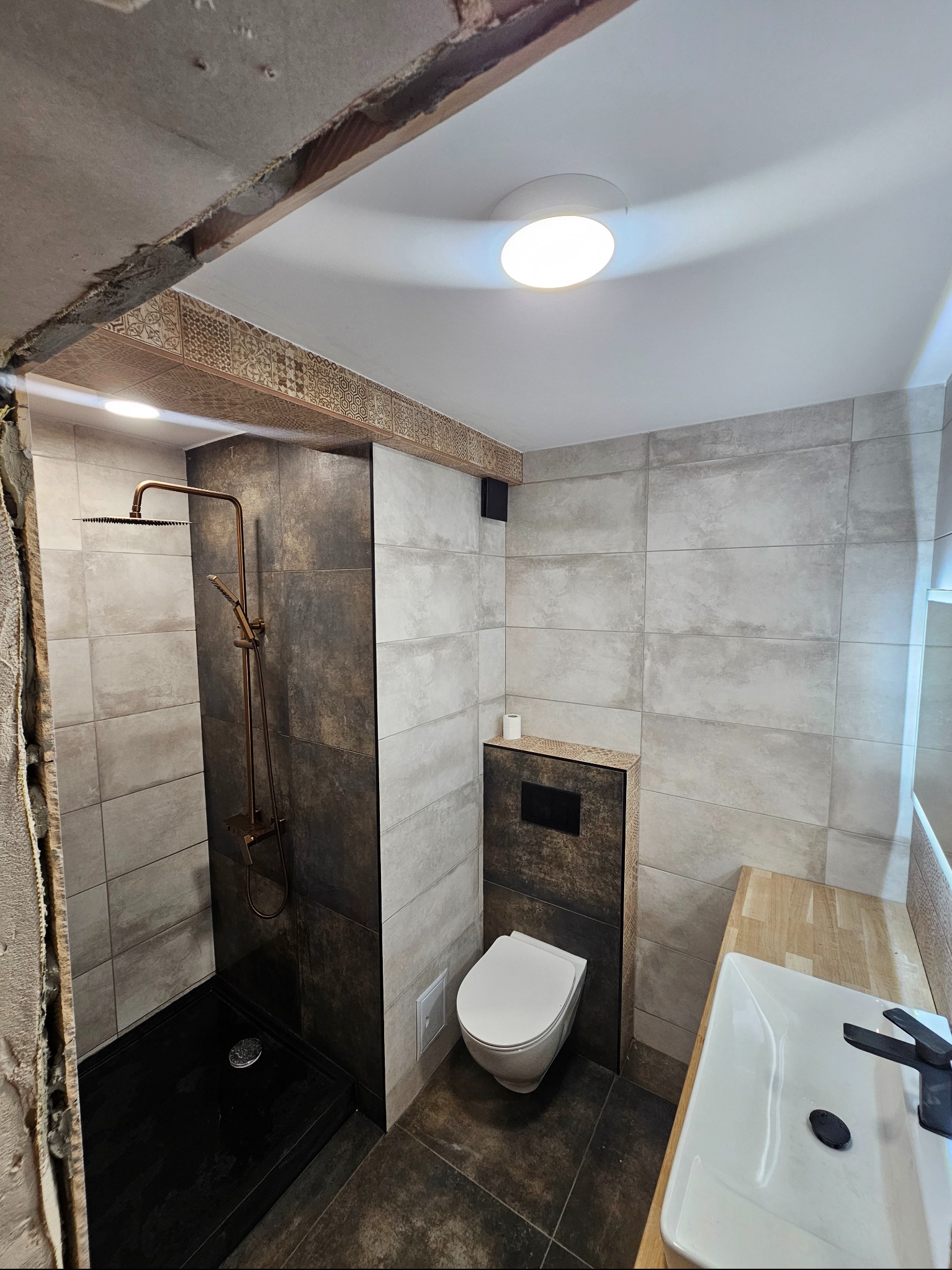 Nowoczesna łazienka w trakcie wykańczania: widoczna kabina prysznicowa z deszczownicą w kolorze miedzi, podwieszana toaleta, umywalka nablatowa na drewnianym blacie oraz fragment odsłoniętej ściany...