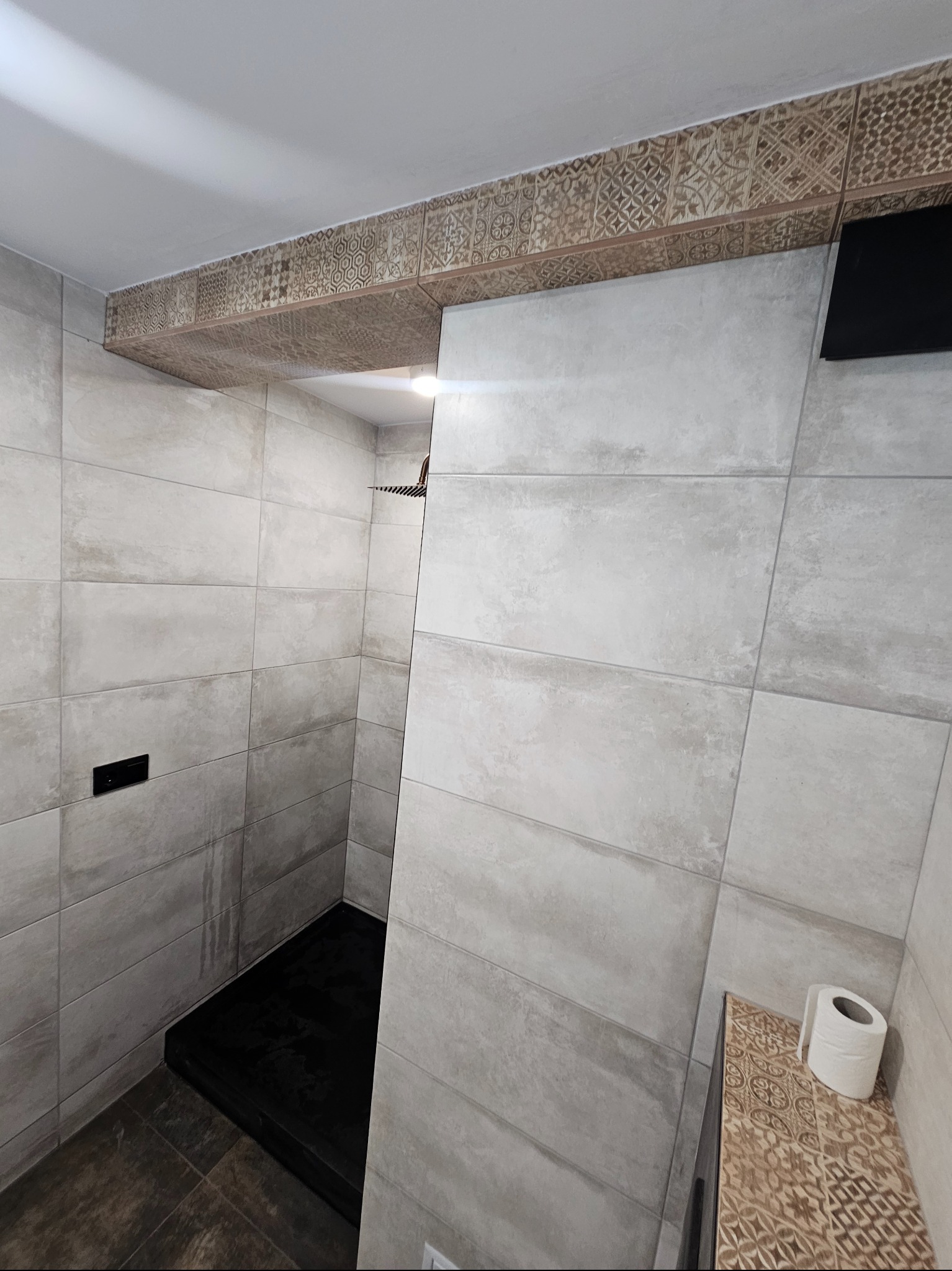 Nowoczesna łazienka z szarymi, prostokątnymi płytkami, czarnym brodzikiem i dekoracyjnym pasem płytek z geometrycznym wzorem pod sufitem oraz rolką papieru toaletowego na półce.