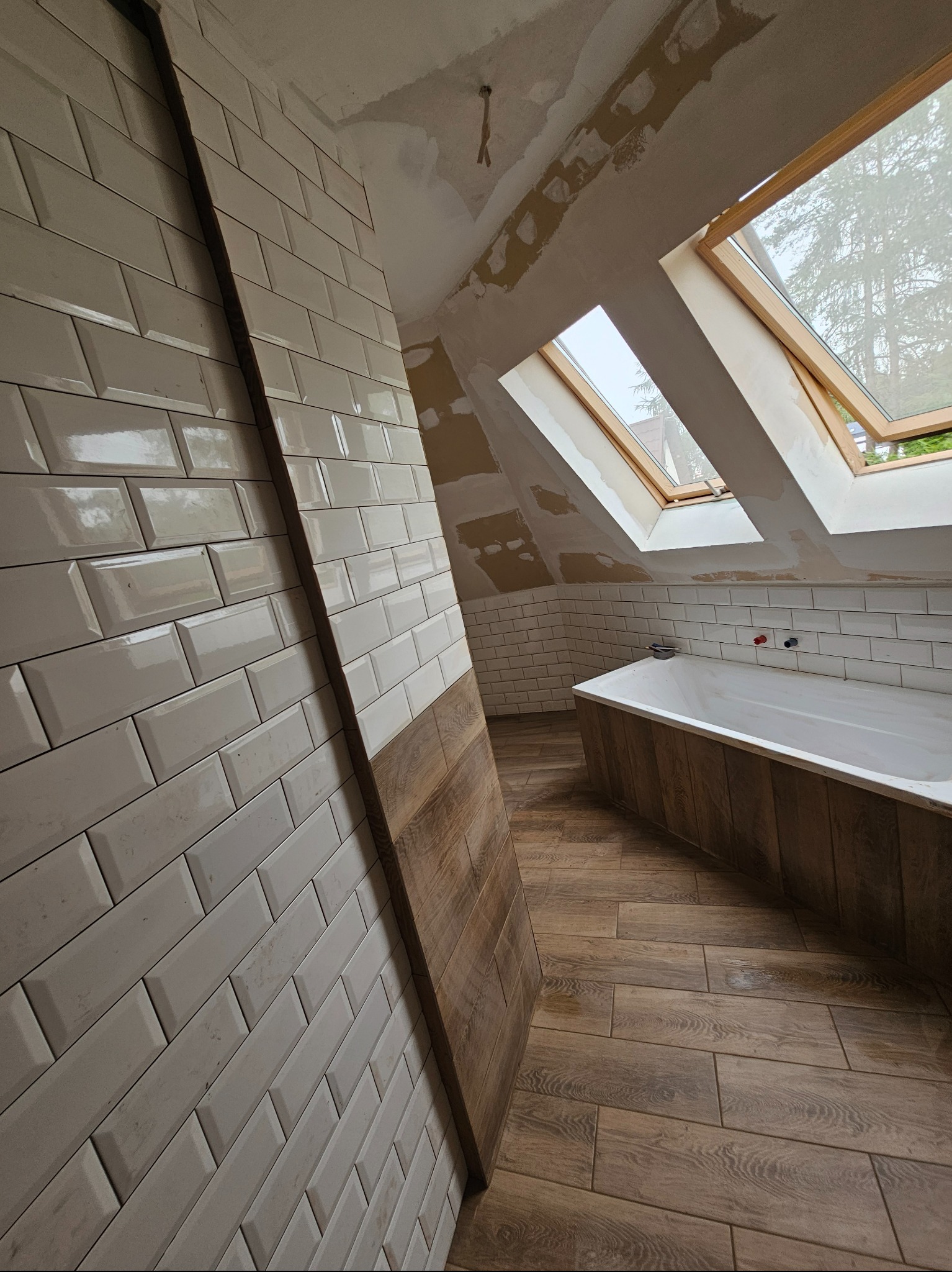 Ukończona łazienka na poddaszu z drewnopodobną podłogą i obudową wanny, białe płytki typu metro na ścianach, dwa okna dachowe z widokiem na drzewa, surowy sufit w trakcie wykończenia.
