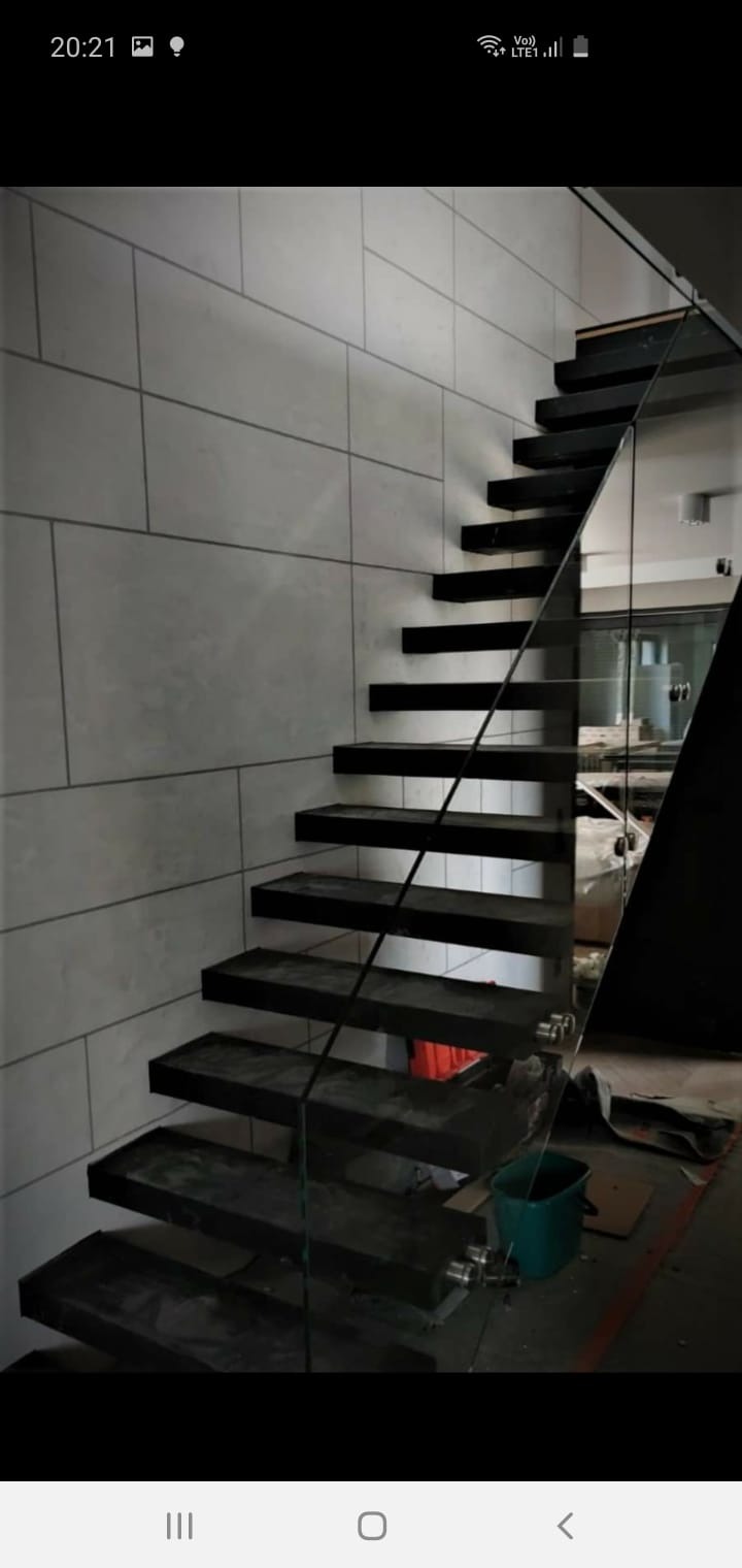 Nowoczesne, wiszące schody z czarnymi stopniami i szklaną balustradą na tle ściany wyłożonej szarymi, prostokątnymi płytami. Widoczne niedokończone wykończenia i wiadro na podłodze.