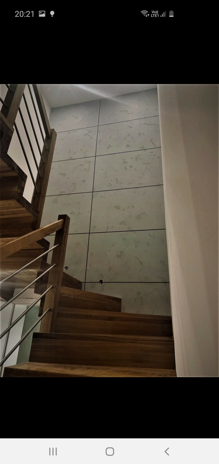 Drewniane schody z metalową balustradą na tle ściany wykończonej płytami imitującymi beton z widocznymi otworami po montażu.