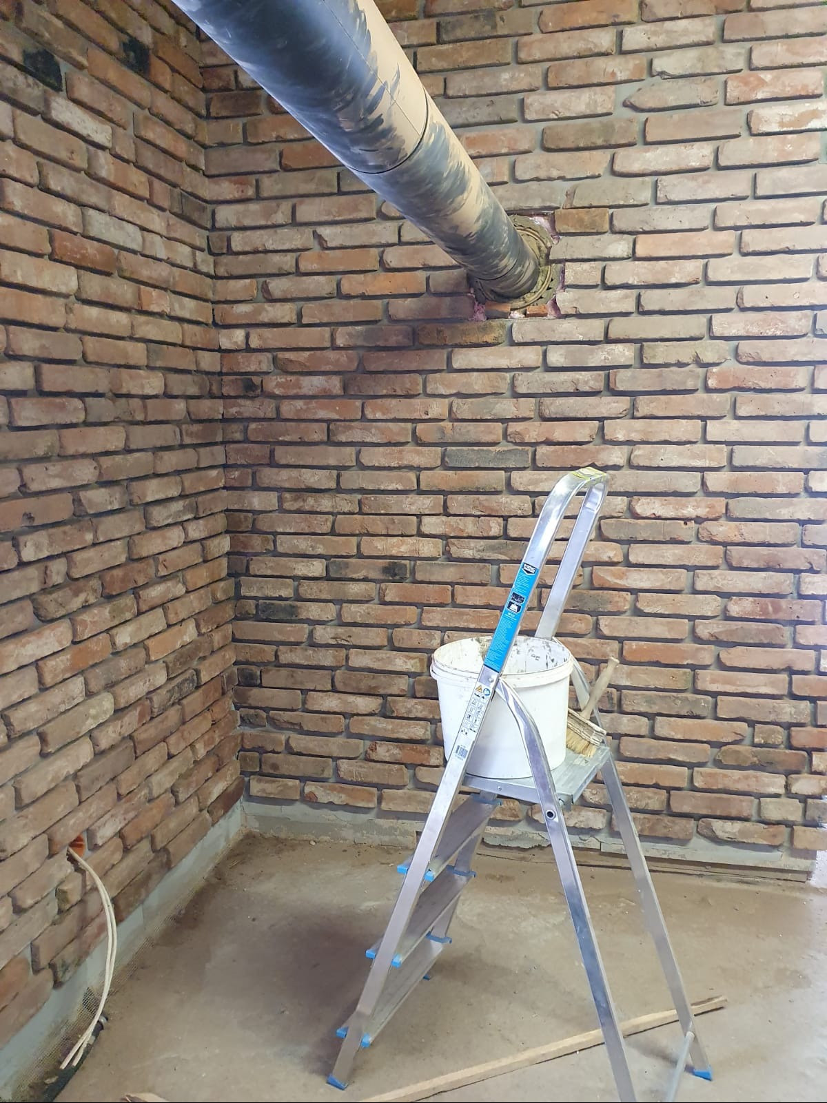 Wnętrze z ceglaną ścianą w trakcie prac wykończeniowych, widoczna drabina z wiadrem farby, wystający rurociąg i kable elektryczne.