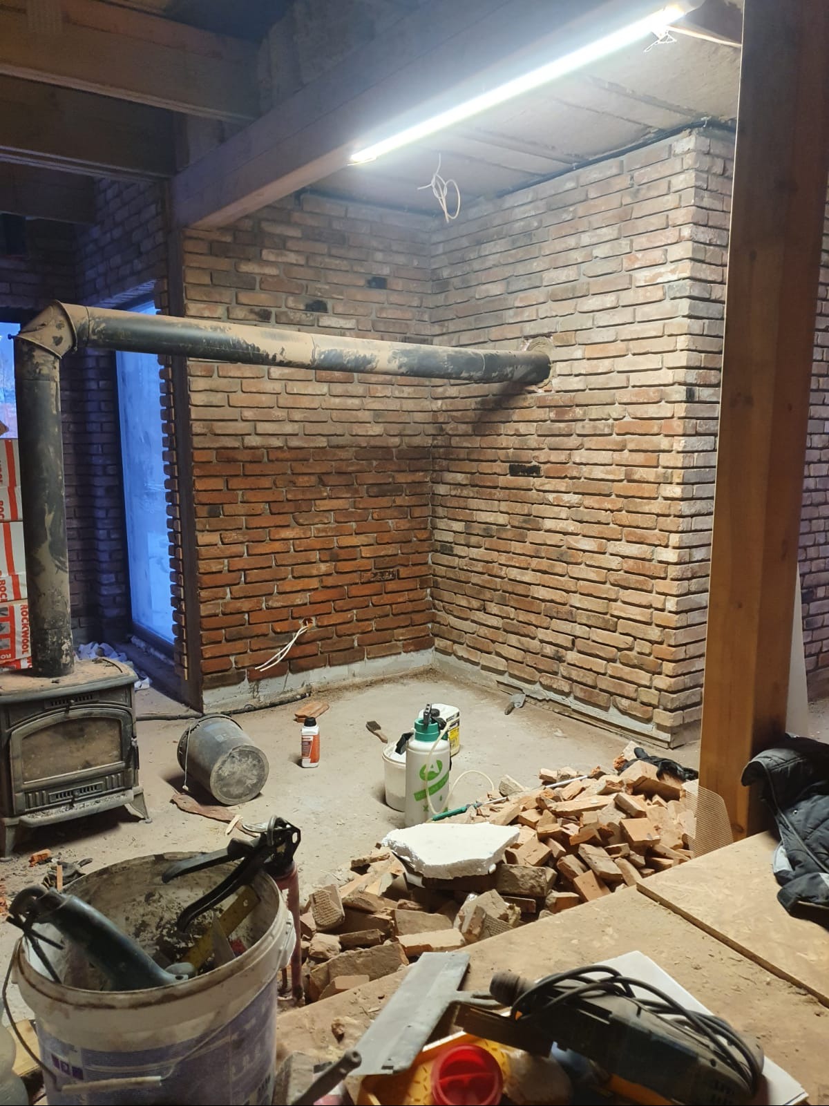 Fragment pomieszczenia w trakcie budowy lub remontu z ceglanymi ścianami, piecem wolnostojącym z rurą odprowadzającą dym, wiadrami, narzędziami i stertą gruzu na podłodze.