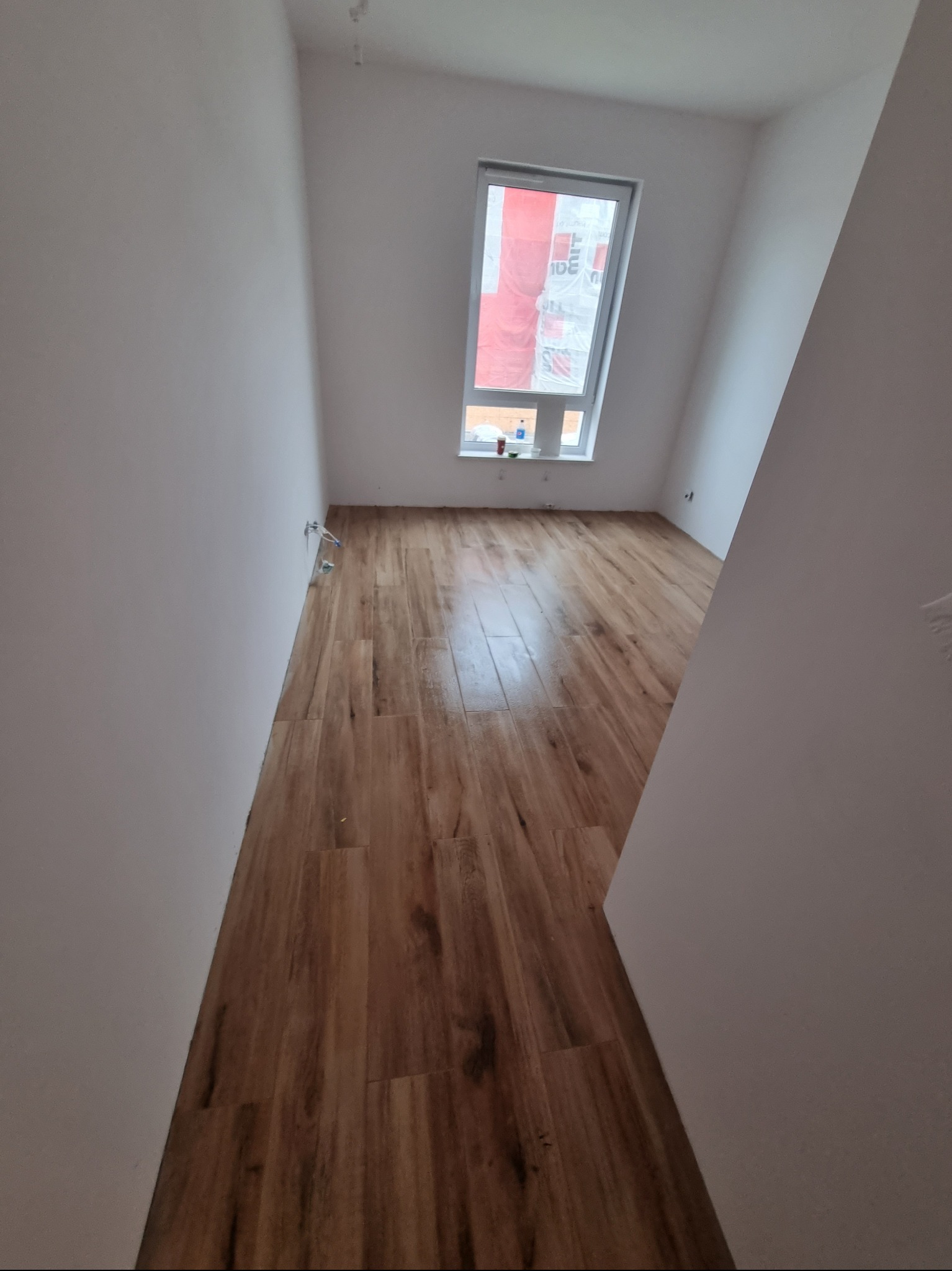 Nowe panele podłogowe imitujące drewno w jasnym pokoju z białymi ścianami i oknem, widoczne przyłącza instalacji elektrycznej.