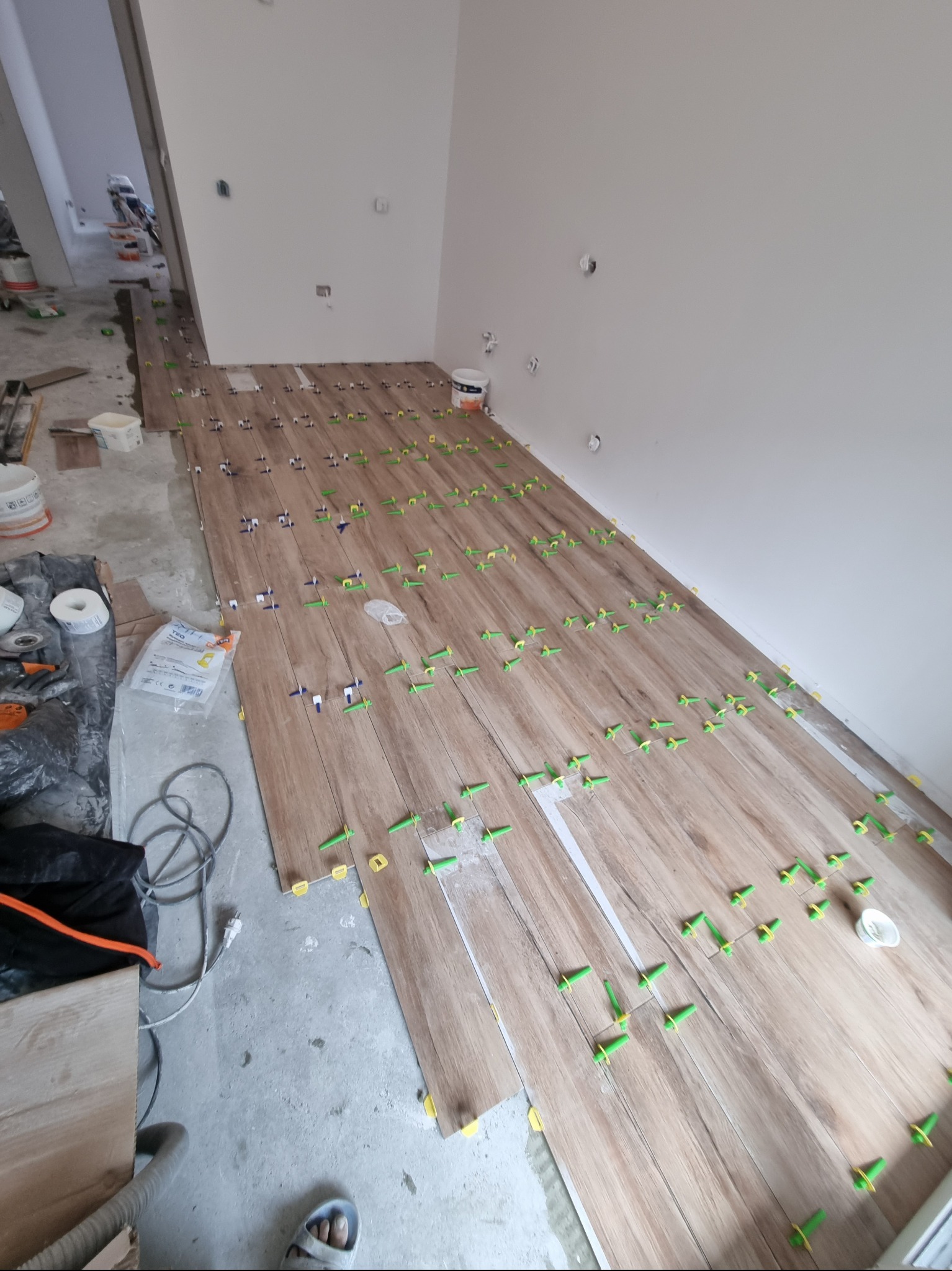 Układanie drewnopodobnych płytek podłogowych z użyciem klipsów poziomujących; widoczny fragment podłogi z przyborami glazurniczymi i stopą w klapku.