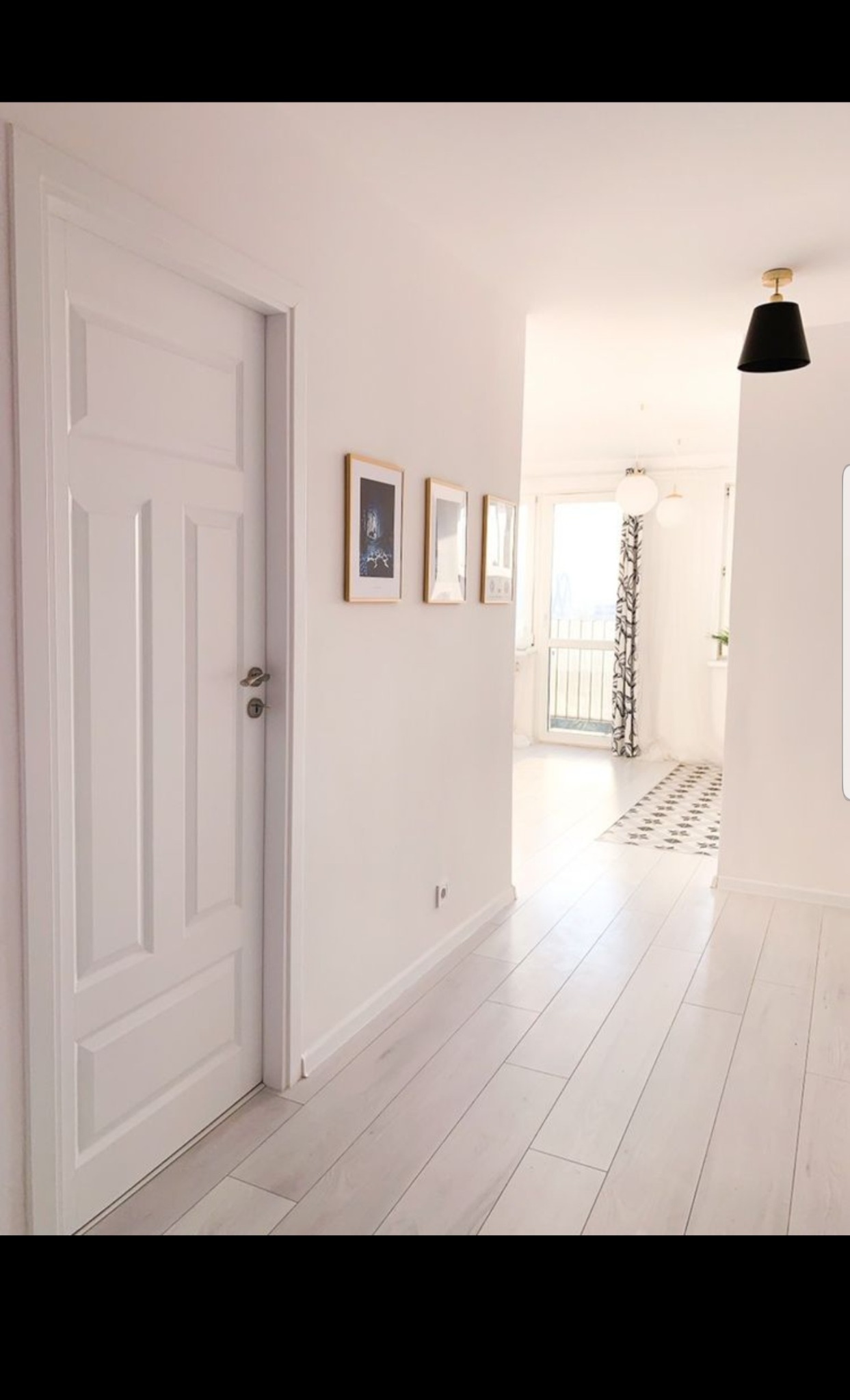 Jasny korytarz z białymi ścianami, drzwiami i podłogą z paneli, ozdobiony trzema obrazami w złotych ramkach i czarną lampą sufitową.