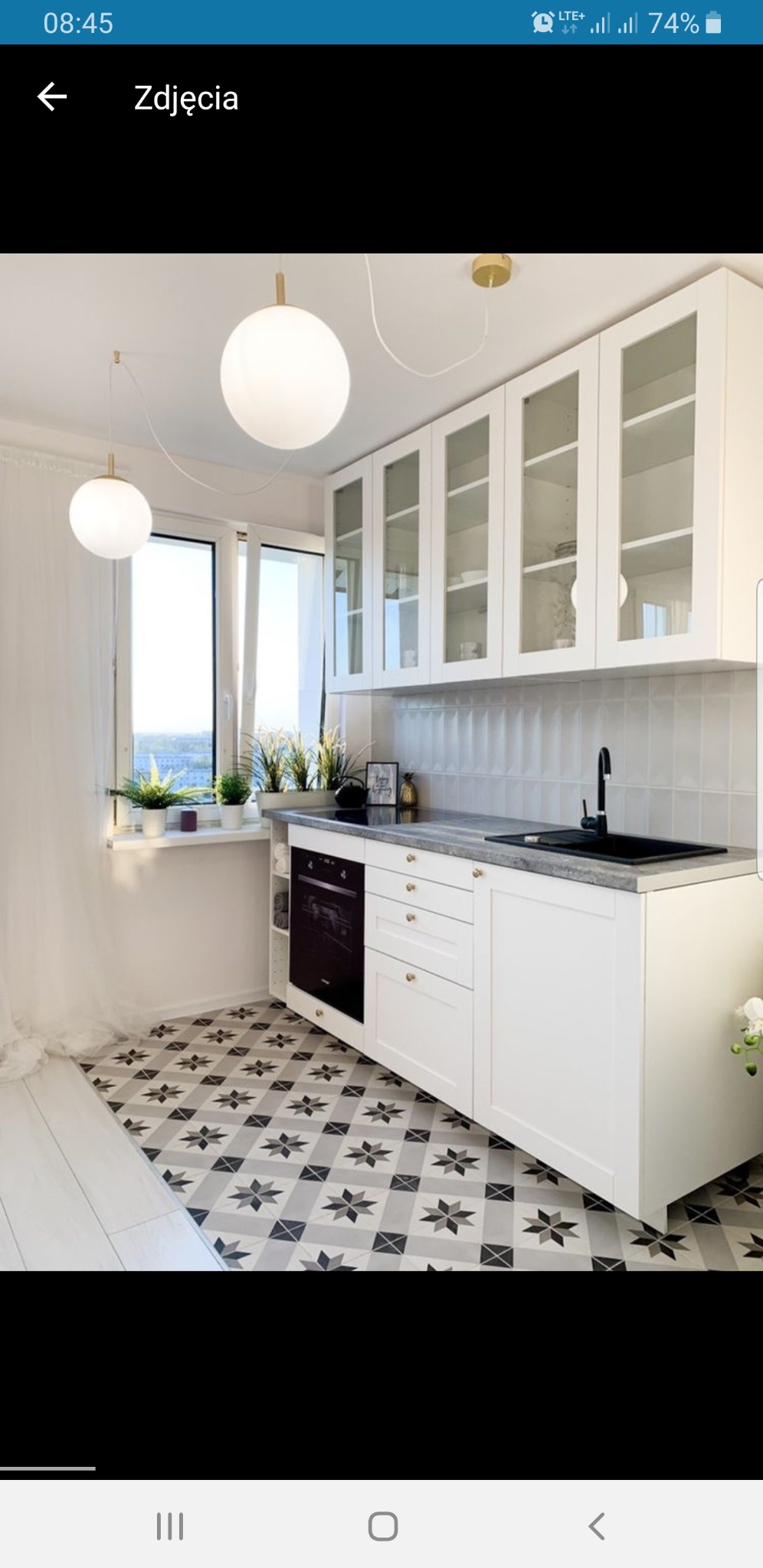 Jasna kuchnia z białymi szafkami z przeszklonymi frontami, czarnym zlewem i płytą, oraz wzorzystą podłogą w gwiazdy, oświetlona kulistymi lampami wiszącymi i światłem wpadającym przez okno...