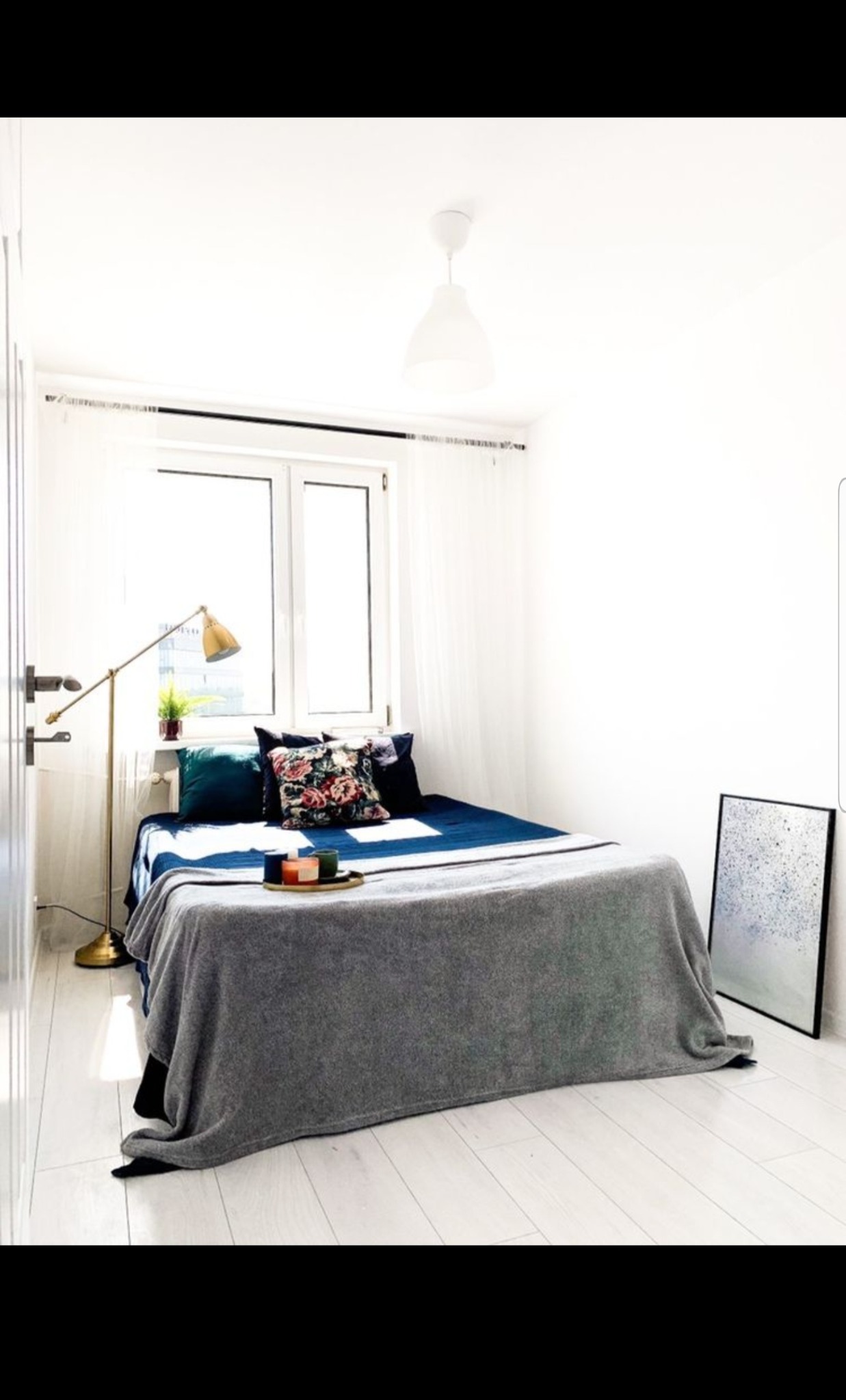 Jasne, minimalistyczne wnętrze sypialni z łóżkiem w niebieskiej pościeli, szarym pledem, lampą podłogową i obrazem opartym o ścianę.