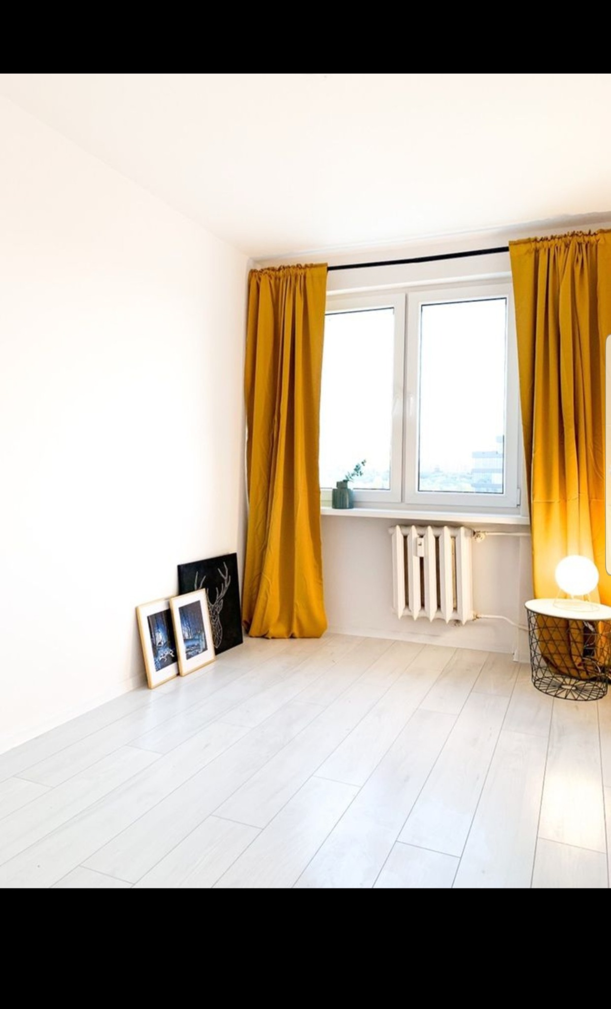 Pusty pokój z białymi panelami podłogowymi, oknem z żółtymi zasłonami, białym grzejnikiem żeberkowym, okrągłym stolikiem z lampką i obrazami opartymi o ścianę.