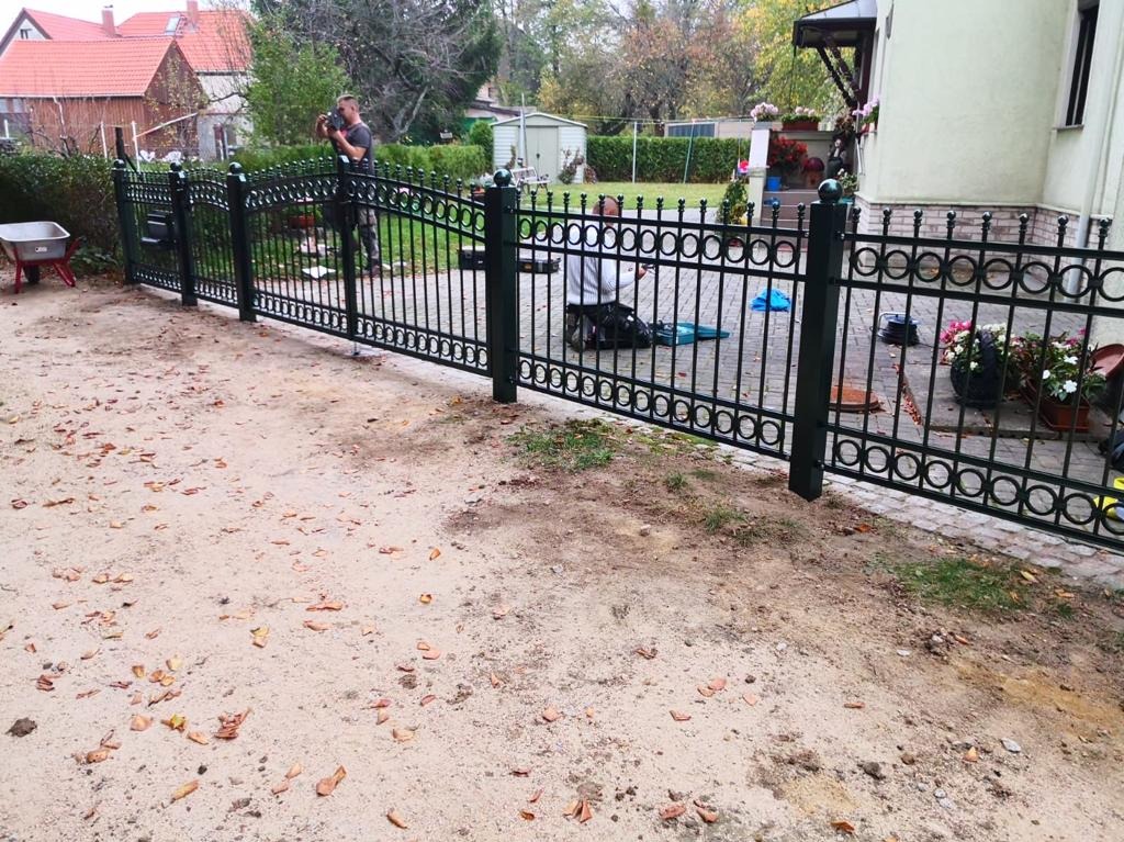 Montaż ozdobnego, metalowego ogrodzenia posesji z okrągłymi elementami dekoracyjnymi; w tle dom jednorodzinny i osoba fotografująca postęp prac.
