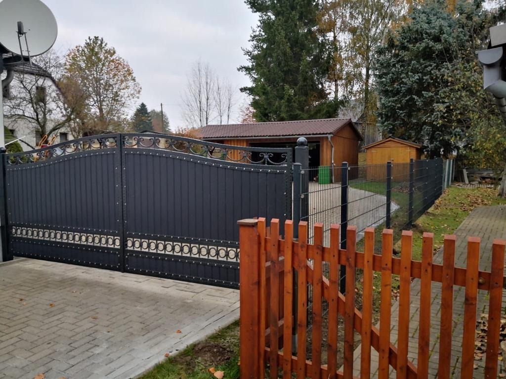 Nowoczesna, czarna brama wjazdowa z dekoracyjnymi elementami, widoczna z perspektywy podjazdu wyłożonego kostką, z fragmentem drewnianego płotu w pierwszym planie i budynkami w tle.