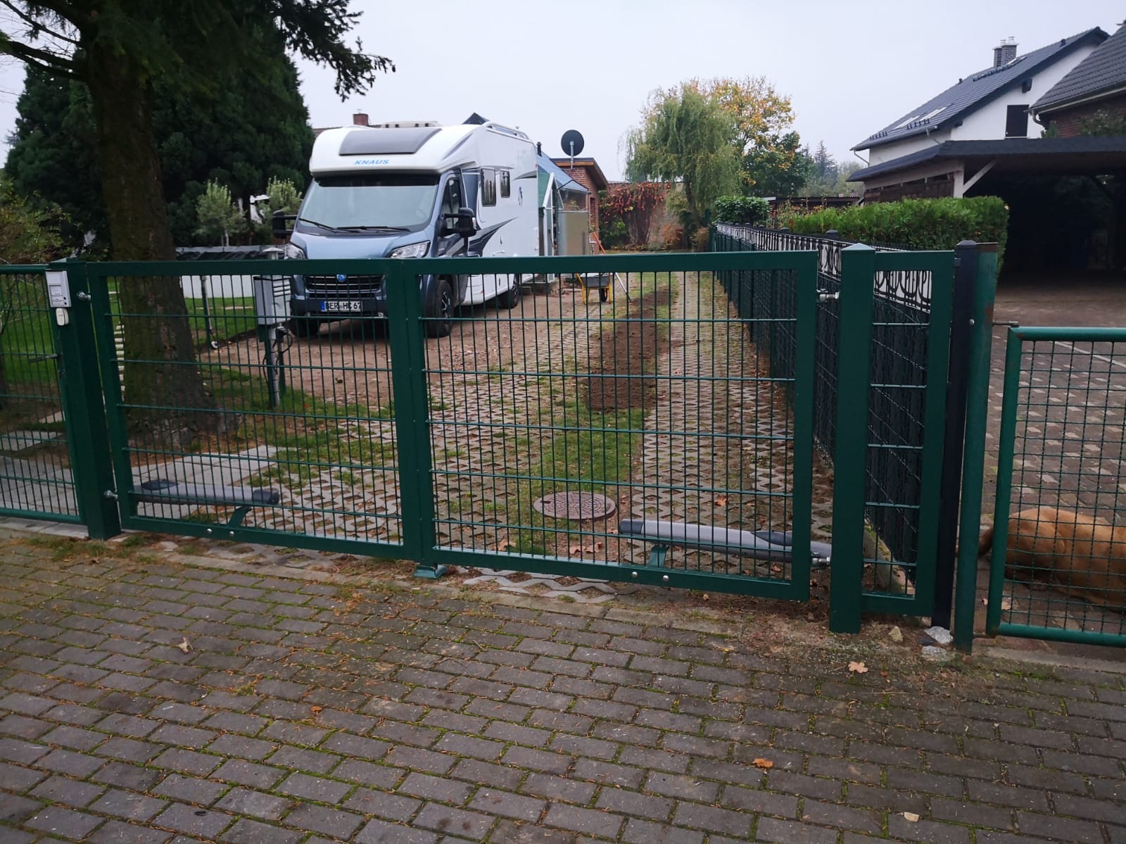 Automatyczna brama wjazdowa z zielonej siatki, widoczny mechanizm otwierania, na podjeździe stoi kamper marki Knaus, w tle dom jednorodzinny.