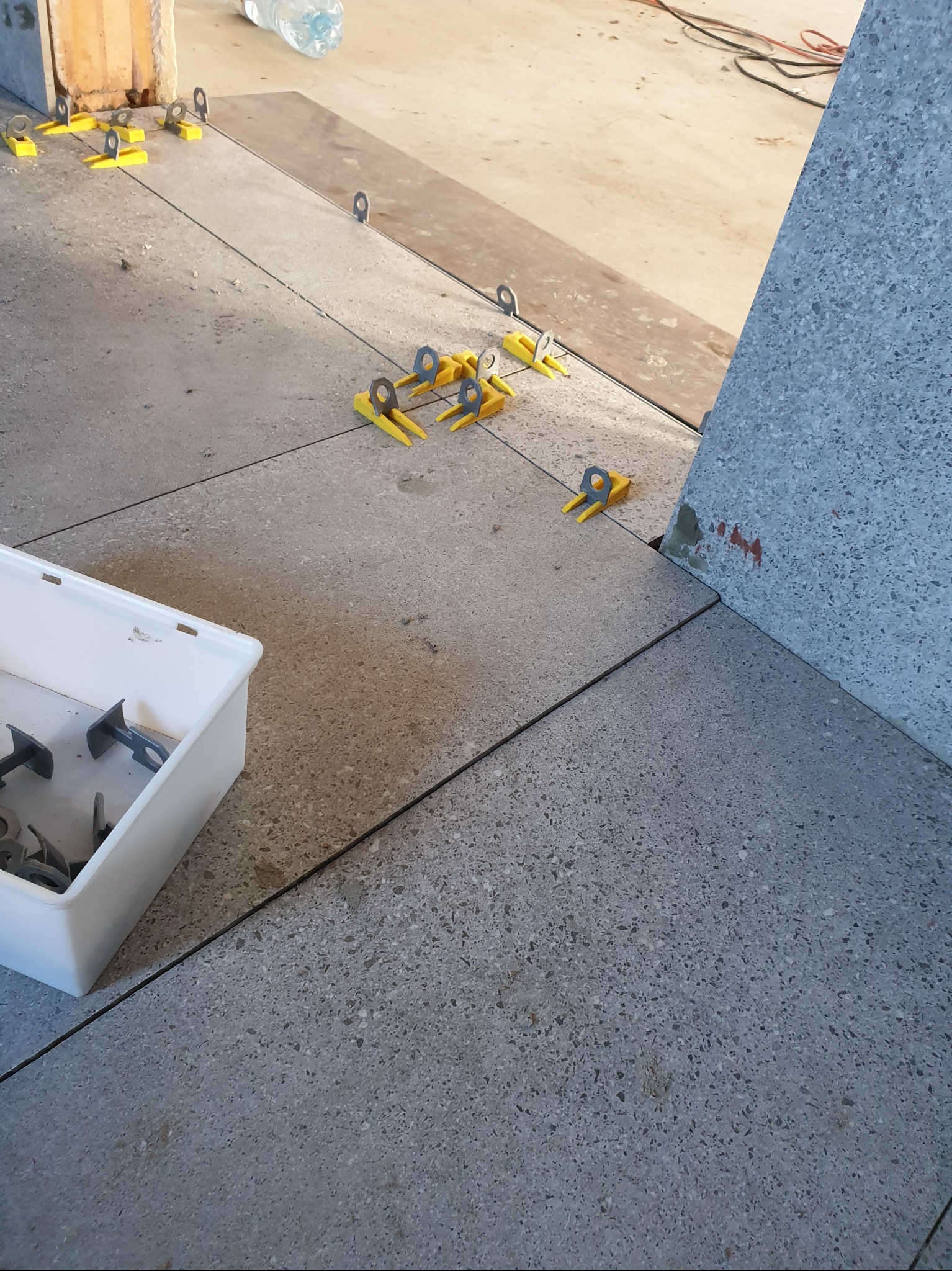 Układanie szarych, nakrapianych płytek podłogowych z użyciem żółtych klinów i metalowych zacisków poziomujących; biały pojemnik z dodatkowymi zaciskami w lewym dolnym rogu.