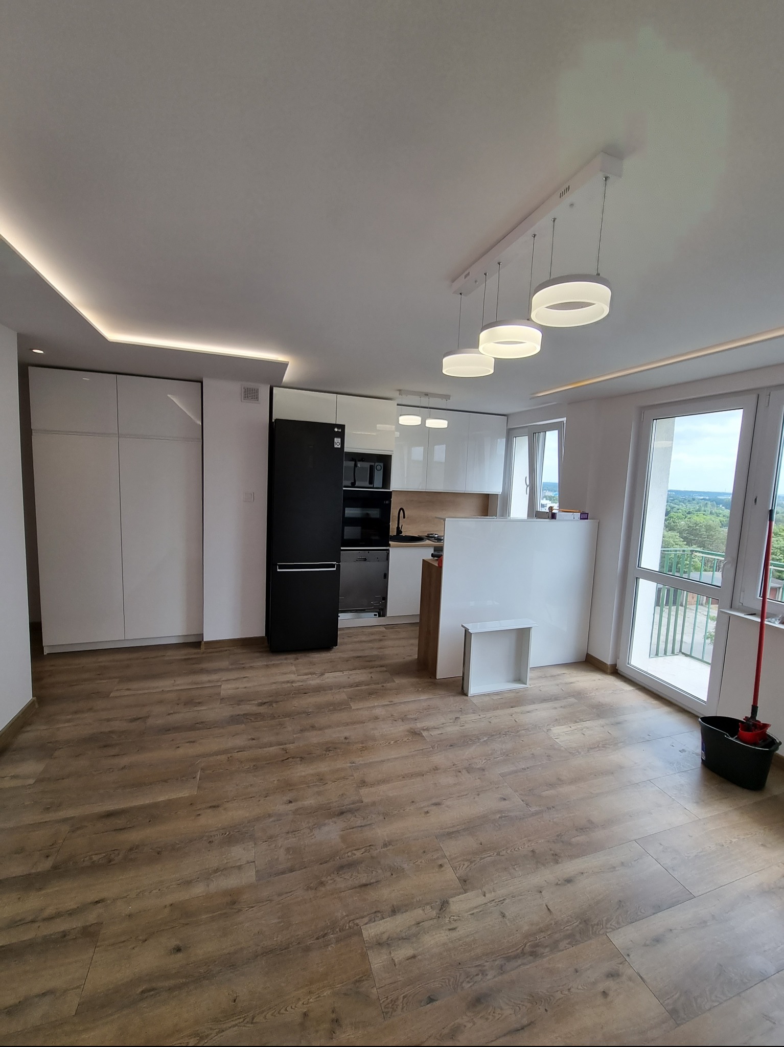 Wykończone wnętrze mieszkania z drewnianą podłogą, białymi szafkami kuchennymi, czarną lodówką LG, nowoczesnym oświetleniem sufitowym i balkonem z widokiem na zieleń.