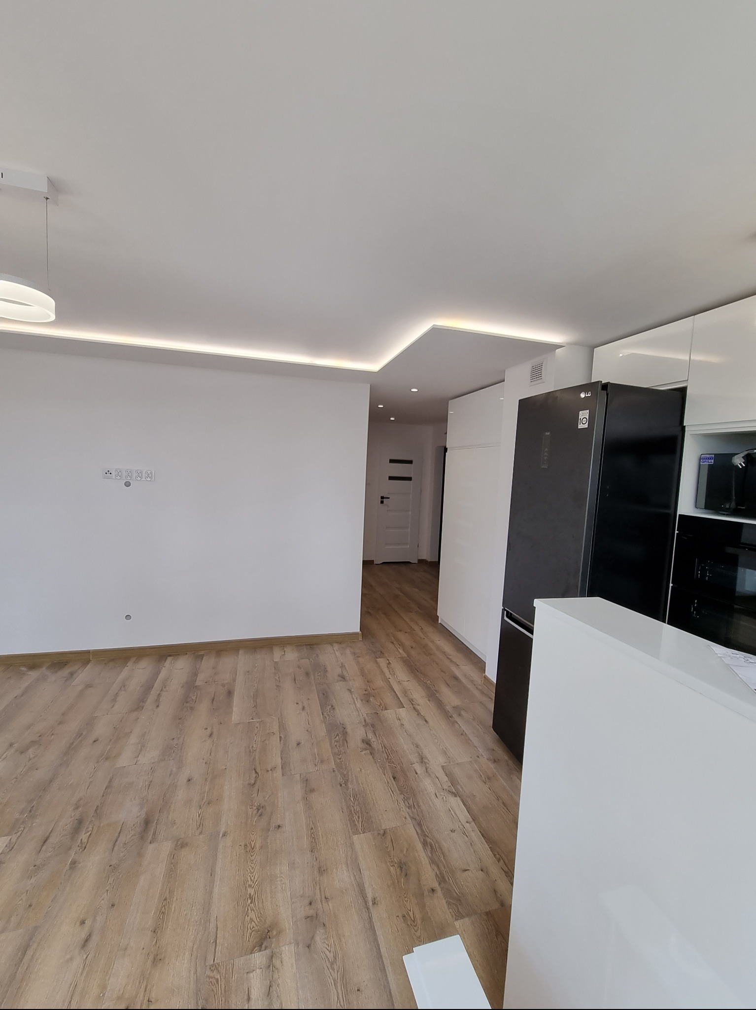 Jasne wnętrze po remoncie: drewnopodobna podłoga, białe ściany z gniazdkami, sufit z oświetleniem LED, zabudowa kuchenna z czarną lodówką LG.