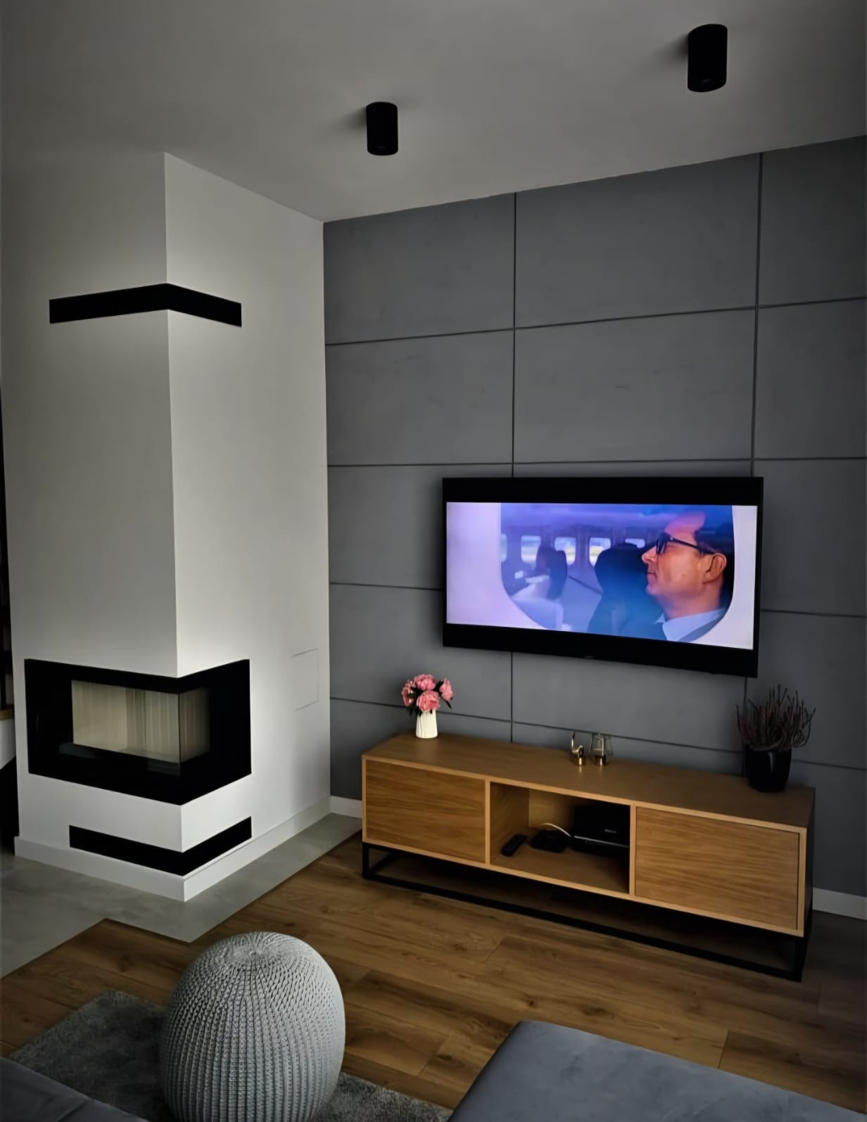 Nowoczesny salon z kominkiem narożnym, szarą ścianą z paneli i drewnianą szafką RTV. Na podłodze panele i pufa. Minimalistyczne oświetlenie sufitowe.