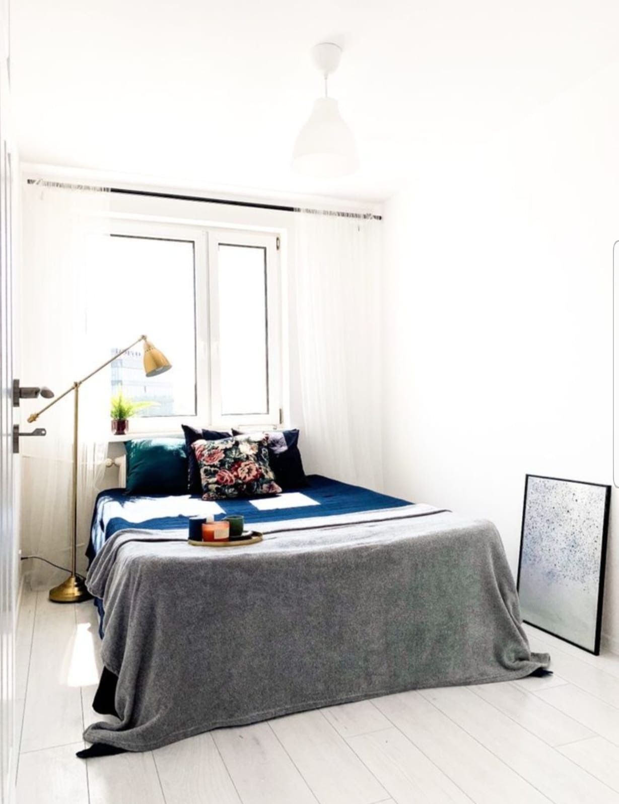 Jasna sypialnia z łóżkiem, lampą podłogową i obrazem. Białe ściany i podłoga, okno z zasłonami. Na łóżku dekoracyjne poduszki i szary pled. Minimalistyczny wystrój.
