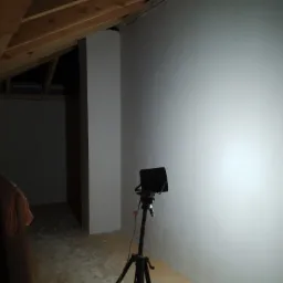 Świeżo otynkowana ściana na poddaszu ze skosami, oświetlona lampą na statywie, widoczne drewniane elementy konstrukcyjne dachu.