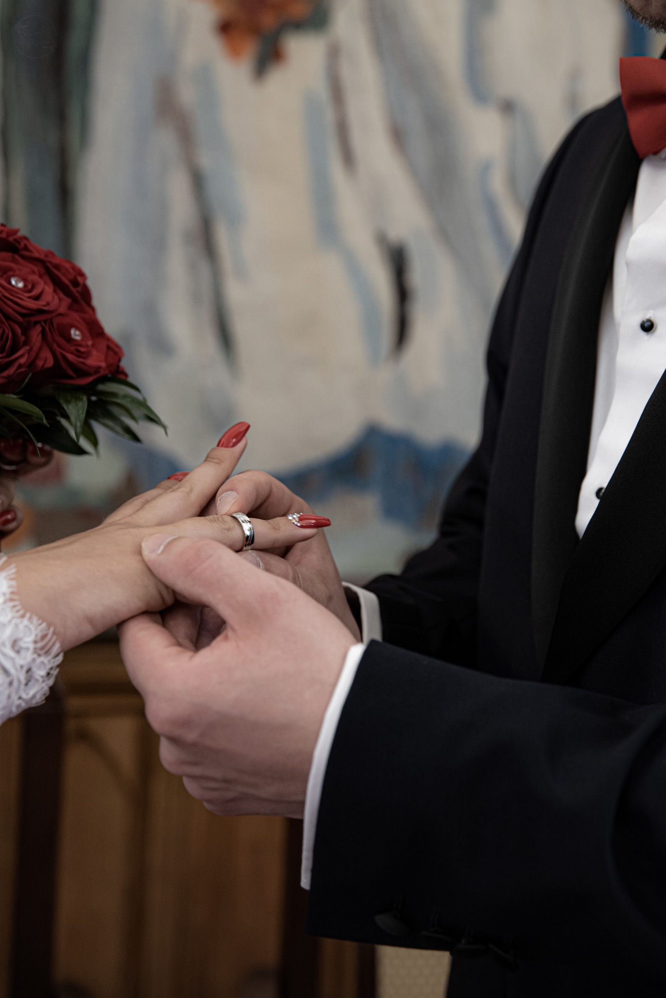 Moment zakładania obrączki podczas ceremonii ślubnej: dłoń panny młodej z czerwonymi paznokciami i bukietem czerwonych róż, dłoń pana młodego w czarnym smokingu z bordową muchą.