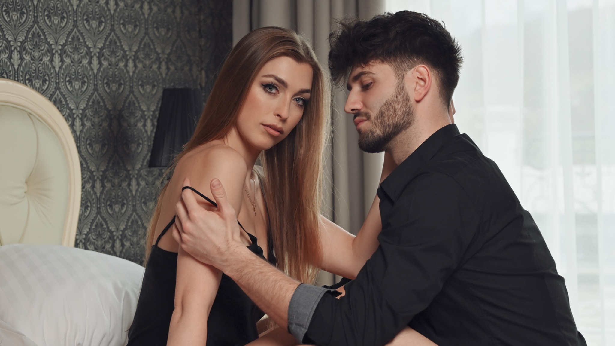 Para w sypialni: kobieta w czarnej bieliźnie, mężczyzna w czarnej koszuli, wzajemne czułe objęcia na łóżku z wzorzystą tapetą w tle.