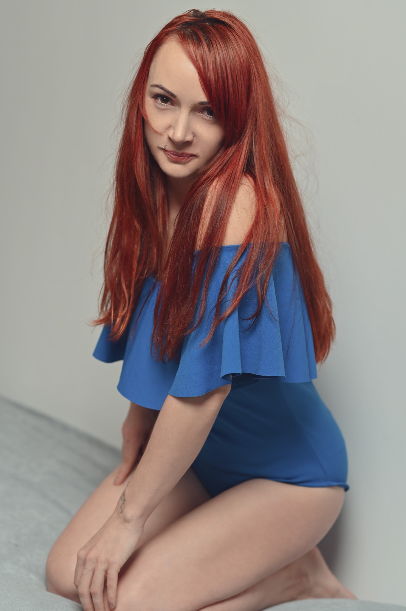Portret kobiety z rudymi włosami w niebieskim stroju z odkrytymi ramionami, z piercingiem w wardze, pozującej na kolanach przy szarej ścianie.