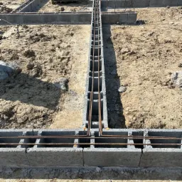 Wykop pod fundamenty z ułożonymi bloczkami betonowymi, zbrojenie pionowe i poziome, widoczne bloczki na palecie w tle.