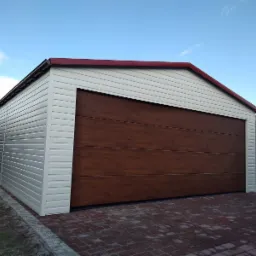 Gotowy garaż z brązową bramą segmentową i białymi ścianami z paneli, czerwone wykończenie dachu, kostka brukowa na podjeździe.