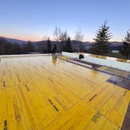 Robert Piwowarczyk Firma ogólno-budowlana - Wylewanie fundamentów pod budowę domu jednorodzinnego na tle górskiego krajobrazu o zmierzchu. Żółte płyty szalunkowe z logo Doka 3S Basic ułożone na zbrojeniu.