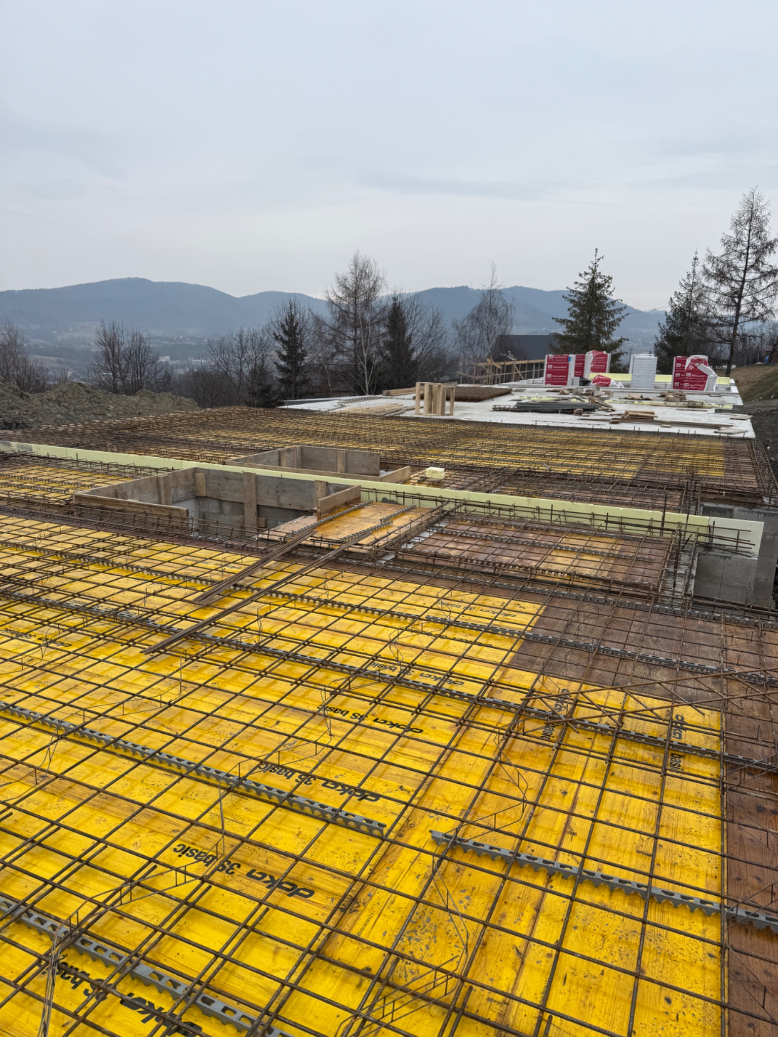 Przygotowanie zbrojenia na żółtej szalunkowej płycie fundamentowej z widocznymi elementami betonowymi i materiałami budowlanymi w tle, krajobraz górski pod pochmurnym niebem.