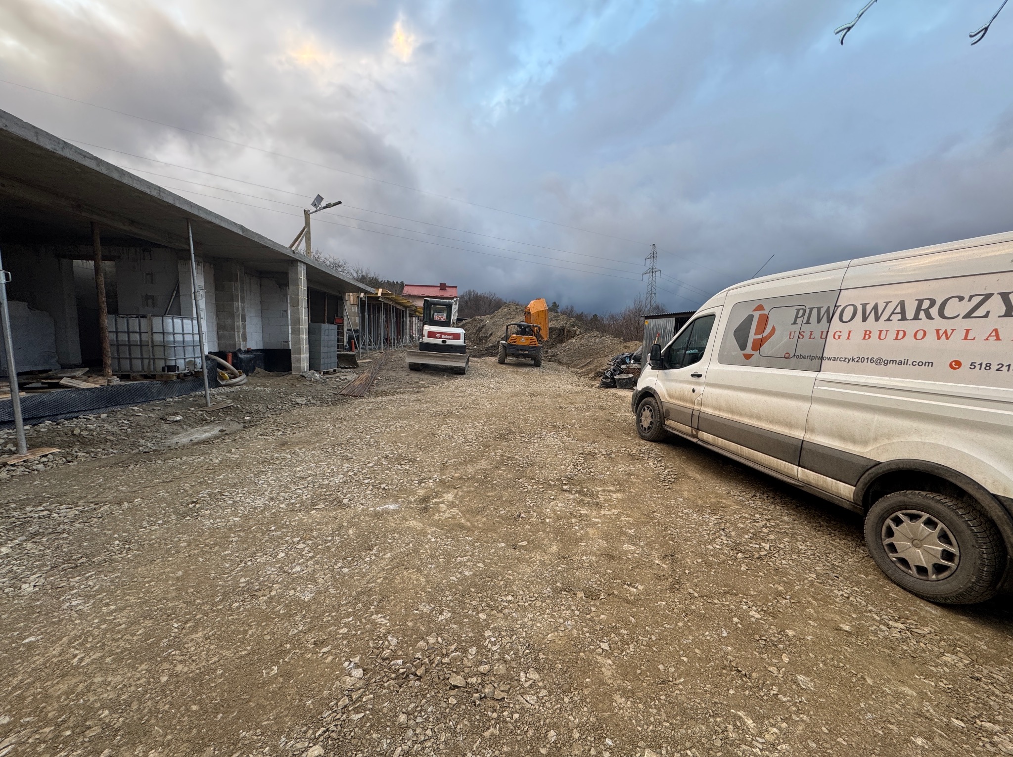 Budowa domu murowanego w trakcie realizacji: widoczny szkielet budynku, dwie maszyny budowlane (koparka i miniładowarka) oraz samochód dostawczy firmy budowlanej Piwowarczyk na placu budowy.